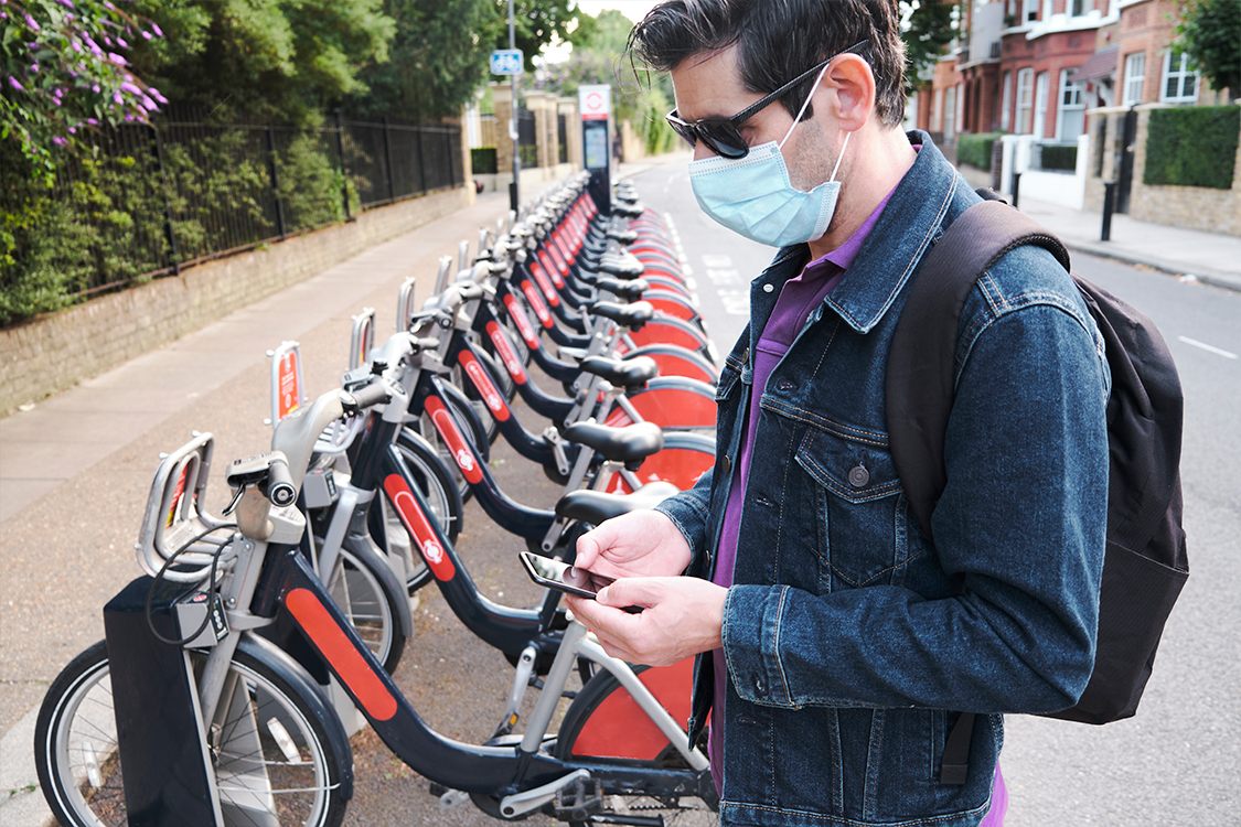 chico con mascarilla consulta su móvil al lado de una estación de aparcamiento de bicicleta pública